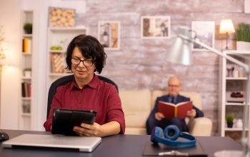 old-lady-her-60s-using-modern-technolgy-cozy-living-space-while-her-husband-reads-background_482257-22297